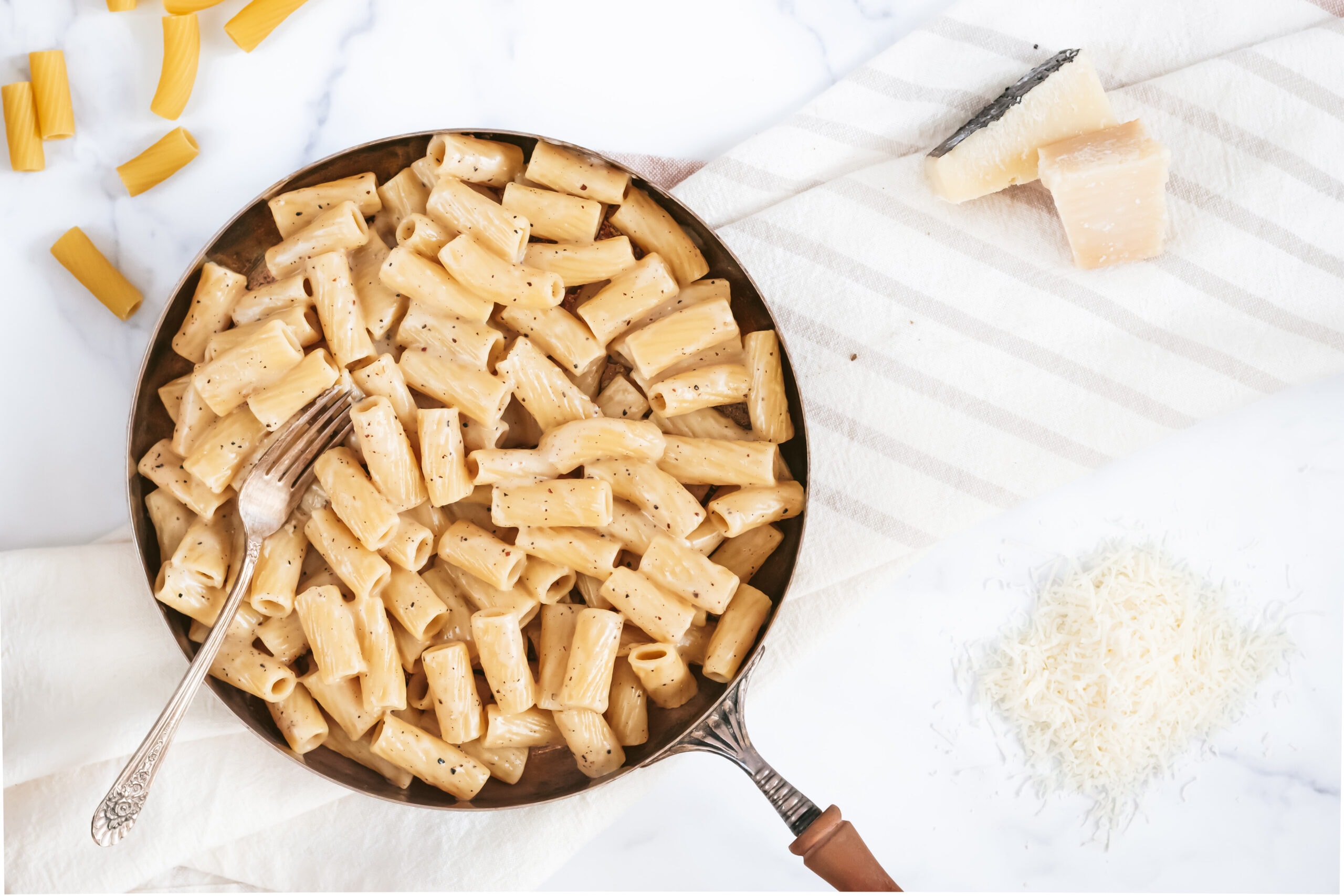 an overhead photo of a skillet of cheesy rigatoni noodles with a fork sticking out to the left. two small bricks of cheese and a pile of shredded cheese.