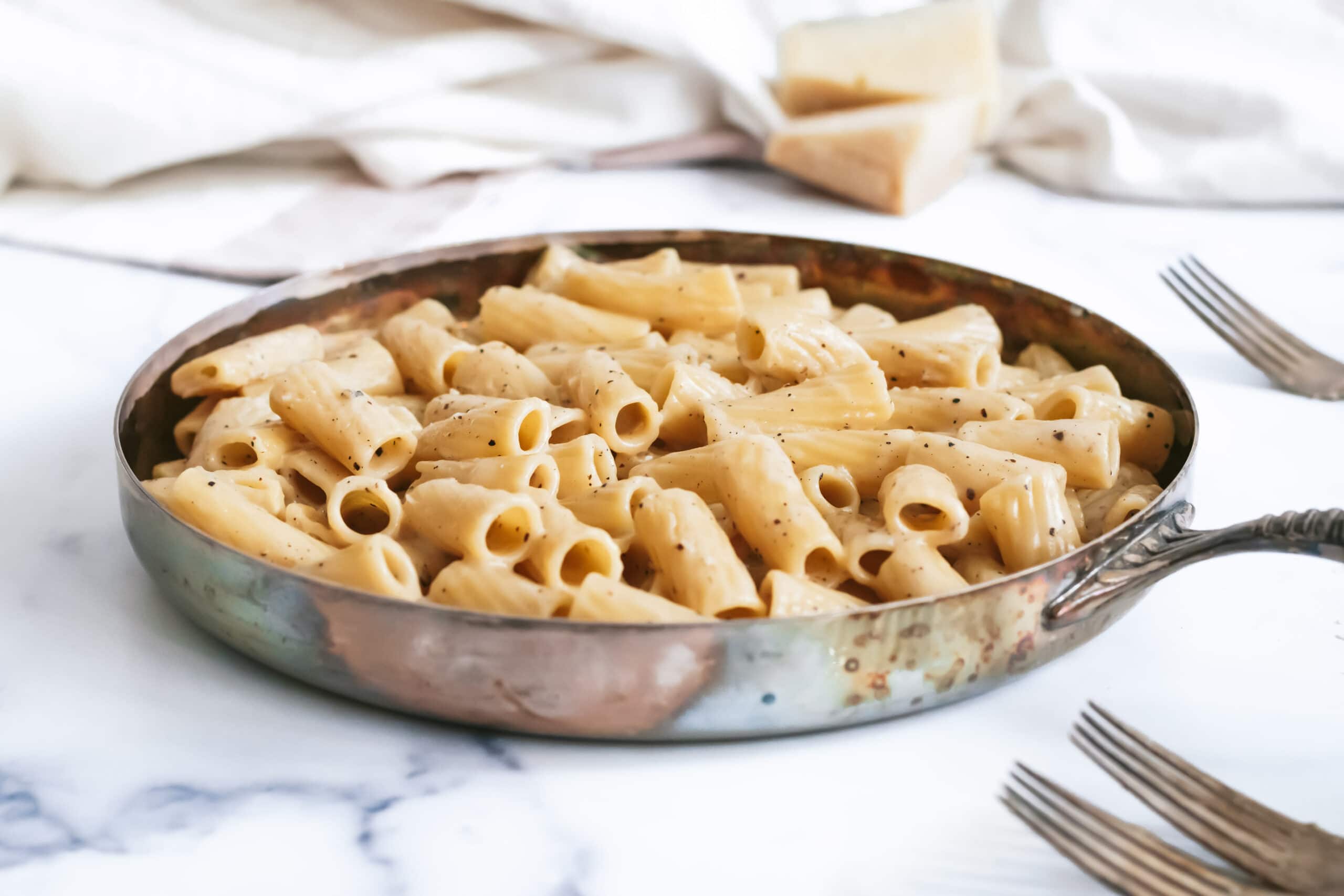 a 45 degree angle shot of a skillet of cheesy cacio e pepe rigatoni. in the background you can see two small wedges of cheese and a linen dish cloth.