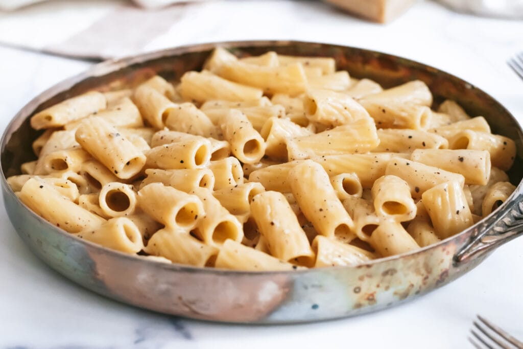 a close up of a skillet with cacio e pepe rigatoni flecked with pepper and shiny melted cheese